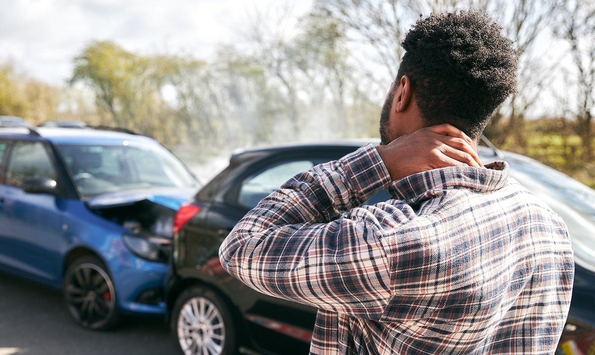 Young man rubbing neck in pain from whiplash injury standing by damaged car after traffic accident